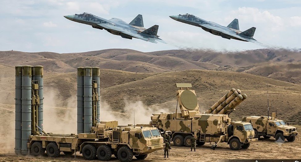 S-500 missile defense system launchers on the ground with two Su-57 stealth fighter jets flying overhead in a clear sky.